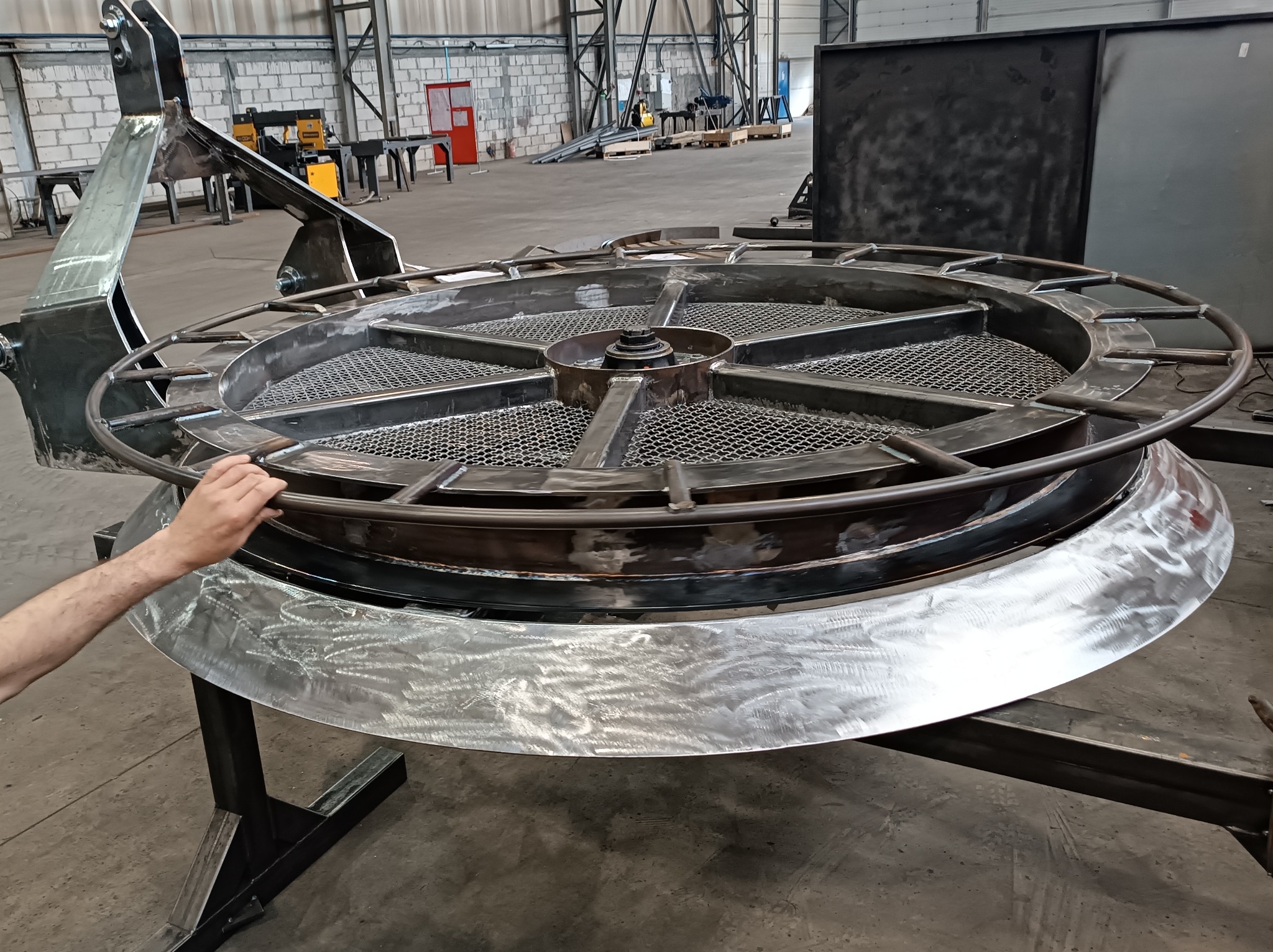 Obrabiany metalowy element konstrukcyjny w kształcie koła z siatkowym wypełnieniem, umieszczony na metalowym stojaku, widoczne ślady obróbki.