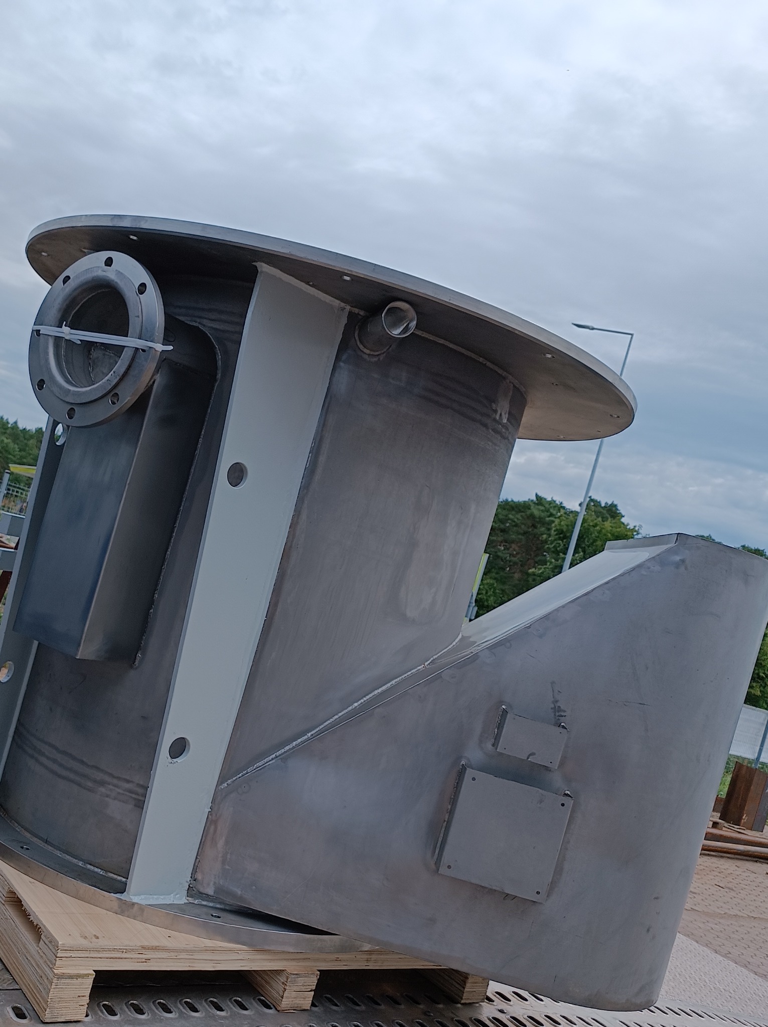 Skomplikowana, spawana konstrukcja ze stali nierdzewnej z kołnierzem i otworami, umieszczona na palecie, sfotografowana z dołu na tle pochmurnego nieba.