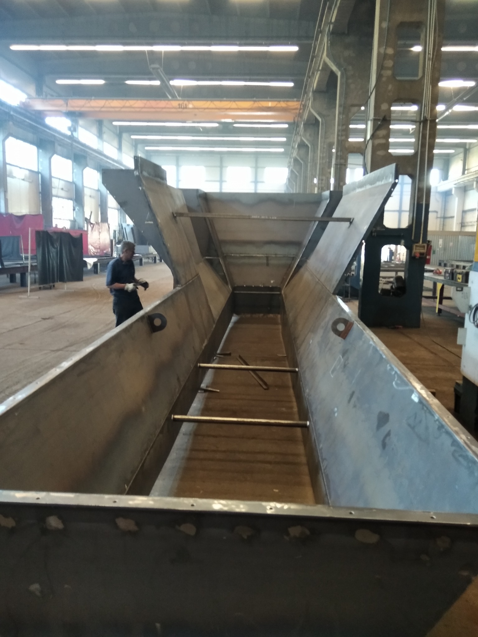 Konstrukcja stalowa w hali produkcyjnej, widoczny spawacz w tle, elementy mocujące i spawane łączenia.