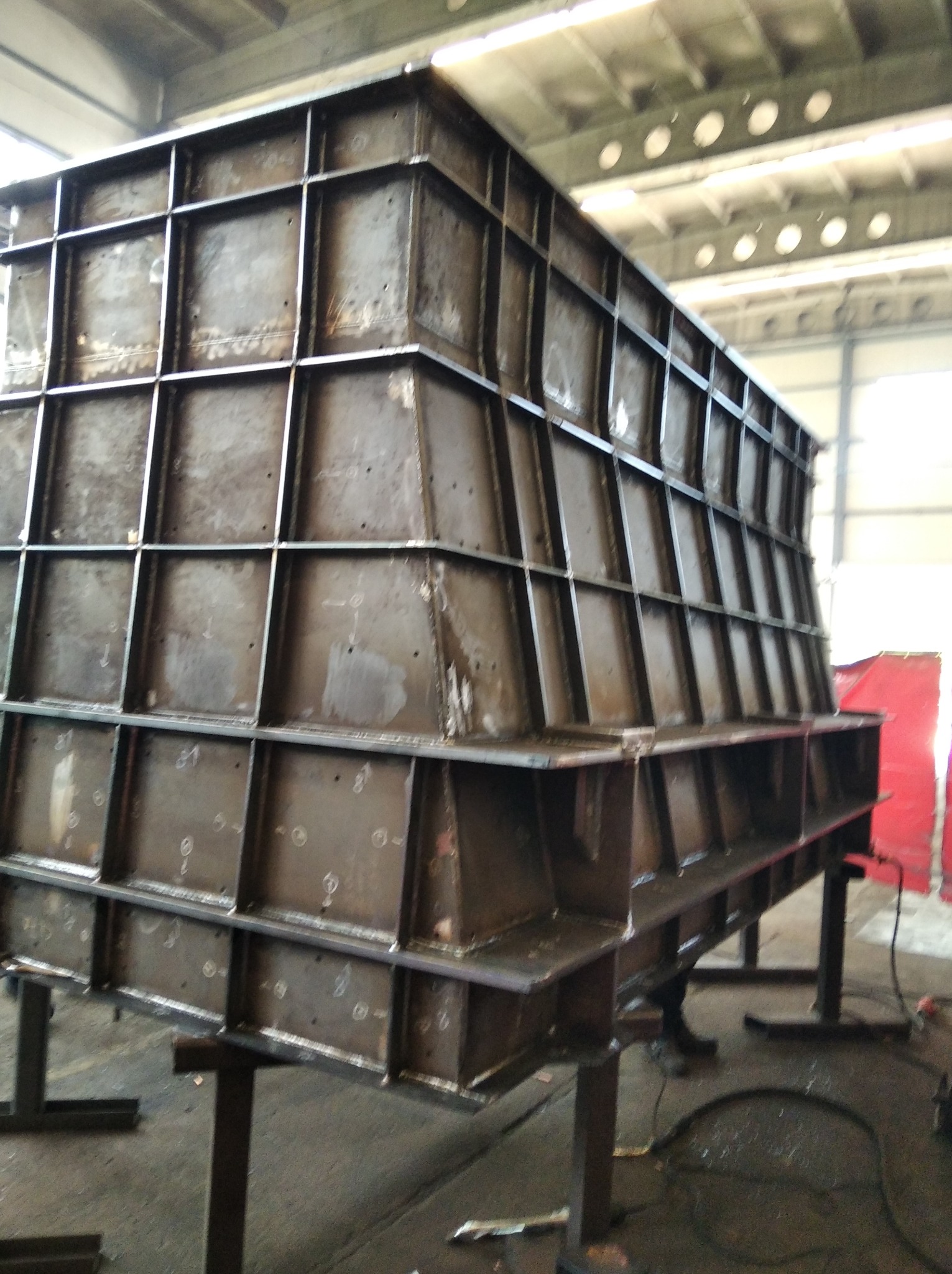 Konstrukcja stalowa zespawana z profili kwadratowych i blach, widoczne ślady spawania i oznaczenia montażowe, ustawiona na podporach w hali produkcyjnej.