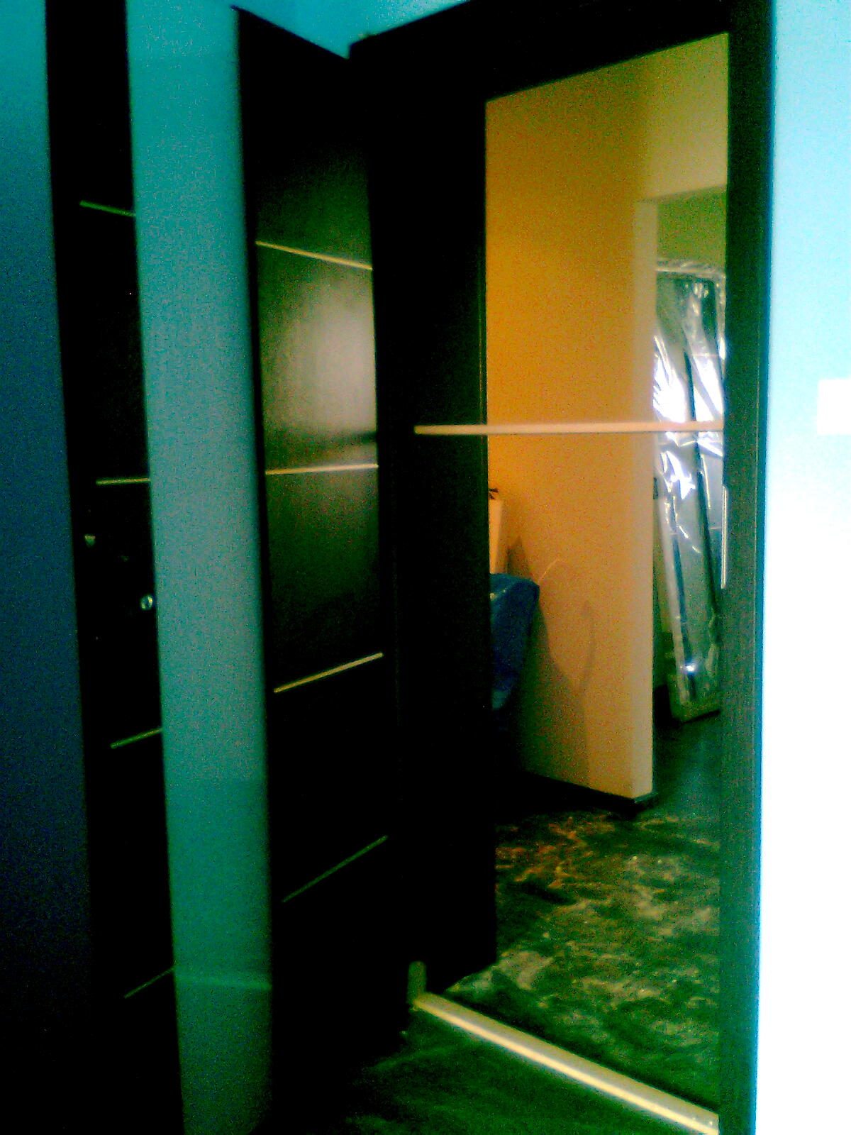 Montaż drzwi wewnętrznych z ciemnego drewna z poziomymi, jasnymi paskami dekoracyjnymi, widok na fragment pomieszczenia w trakcie prac wykończeniowych, zabezpieczone folią elementy.