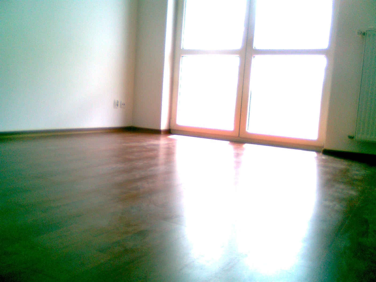 Wykończony pokój z drewnianą podłogą, świeżo po lakierowaniu, z dużym oknem balkonowym w tle, wpadające światło odbija się od błyszczącej powierzchni podłogi.