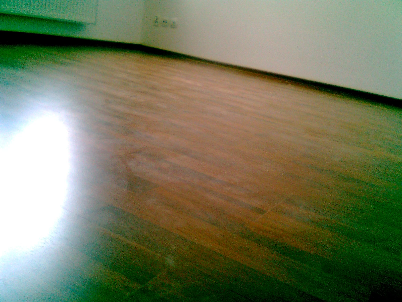 Ułożone brązowe panele podłogowe z widoczną listwą przypodłogową i gniazdkami elektrycznymi w tle, oświetlone ostrym światłem.