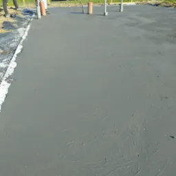 Świeżo wylana, szara płyta betonowa z widocznymi śladami po zacieraczce, obok pracownik z maszyną do zacierania betonu, wystające rury instalacyjne, w tle ogrodzenie i budynek.