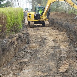 A.L Budowy - Żółta koparka CAT 308 na gąsienicach w trakcie wykopu rowu w ziemi, widoczny operator w żółtej kamizelce, w tle drugi mniejszy sprzęt.