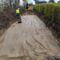 A.L Budowy - Przygotowanie terenu pod budowę: ubijanie ziemi zagęszczarką spalinową i równanie terenu koparką, widoczne ślady opon i świeżo wykopana ziemia po bokach.