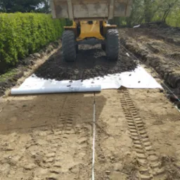 Żółta ładowarka z pełną łyżką ziemi na przygotowanym podłożu z geowłókniny, widoczne ślady opon na piasku, w tle zielony żywopłot.