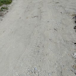 A.L Budowy - Niejednolita, szara nawierzchnia drogi gruntowej z widocznymi śladami opon samochodowych i luźnymi kamieniami, częściowo porośnięta trawą, w tle zaparkowane samochody.