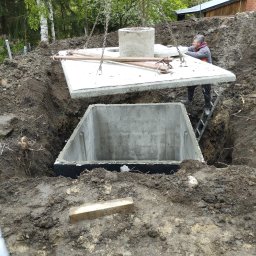 A.L Budowy - Montaż betonowego szamba: Robotnik na drabinie pomaga w opuszczeniu betonowej pokrywy na zbiornik, widoczne łańcuchy dźwigu, wykop ziemny i otaczająca zieleń.