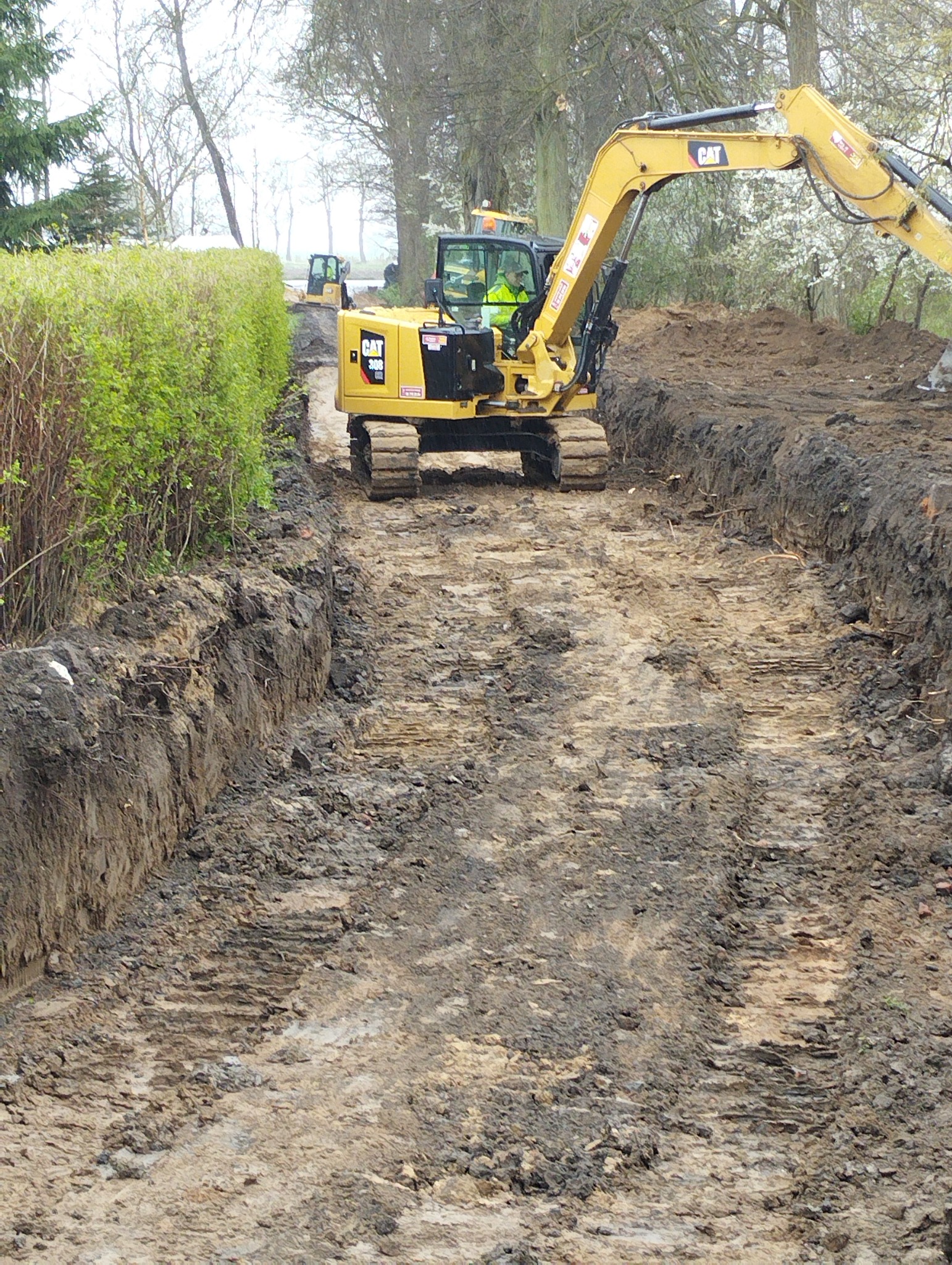 Żółta koparka CAT 308 na gąsienicach w trakcie wykopu rowu w ziemi, widoczny operator w żółtej kamizelce, w tle drugi mniejszy sprzęt.