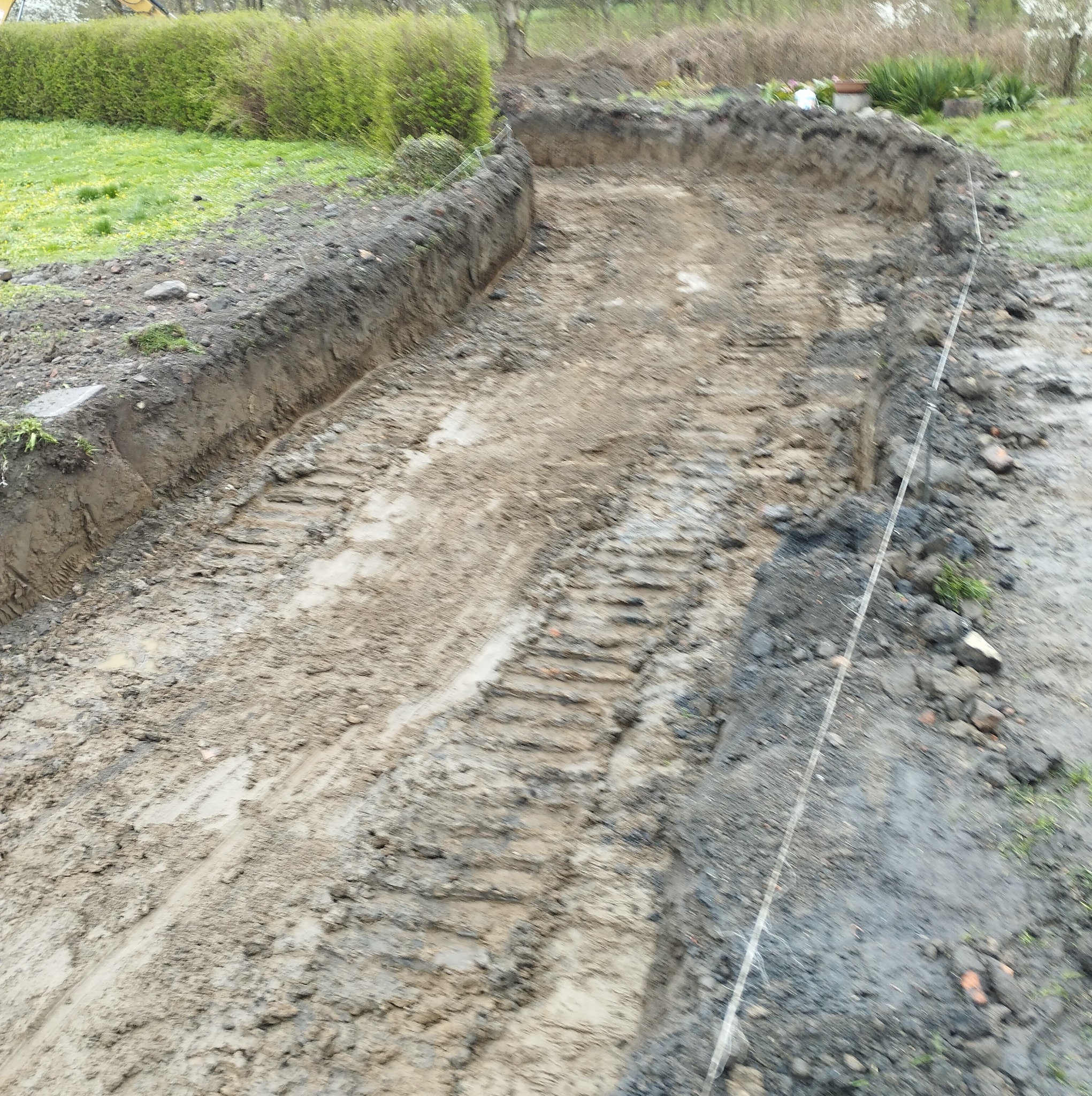 Wykopany rów w ziemi z widocznymi śladami opon maszyny budowlanej, obok trawa i krzewy, wyznaczony sznurek.