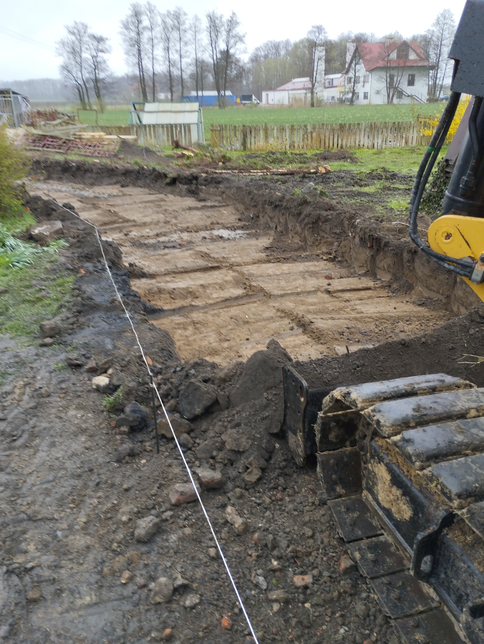 Wykop liniowy w ziemi, wyznaczony sznurkiem, obok gąsienic koparki, w tle zabudowania gospodarcze i dom.
