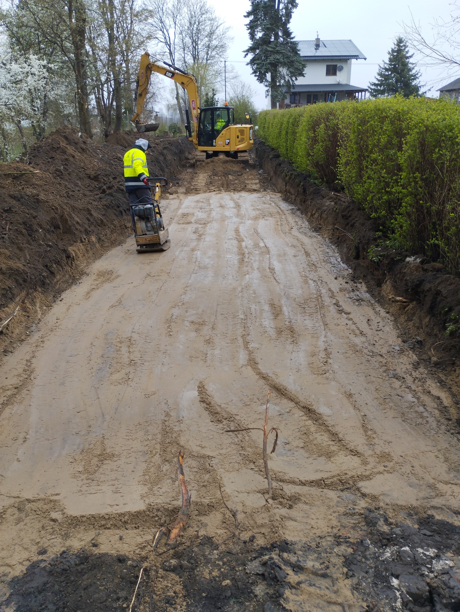 Przygotowanie terenu pod budowę: ubijanie ziemi zagęszczarką spalinową i równanie terenu koparką, widoczne ślady opon i świeżo wykopana ziemia po bokach.