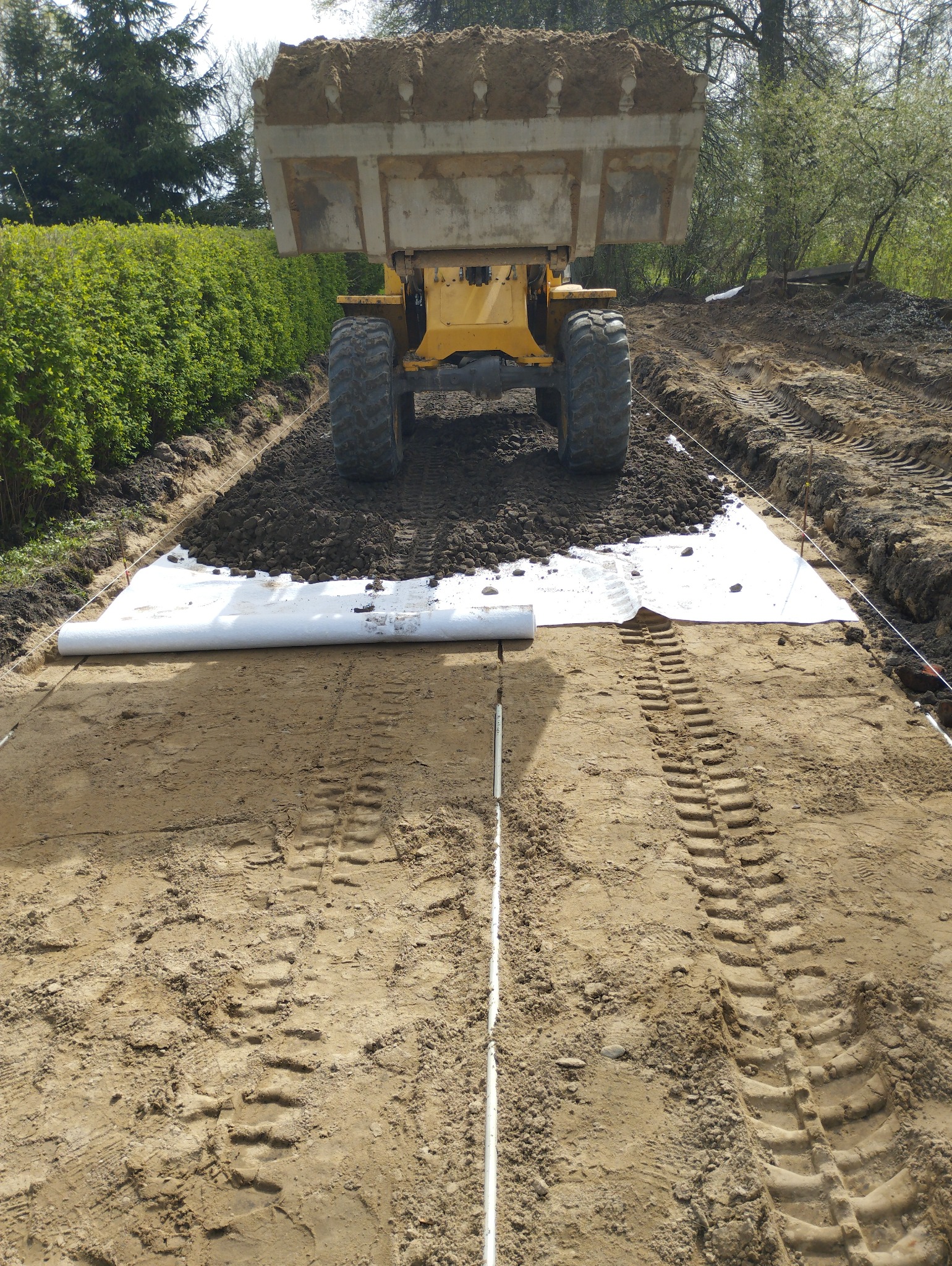 Żółta ładowarka z pełną łyżką ziemi na przygotowanym podłożu z geowłókniny, widoczne ślady opon na piasku, w tle zielony żywopłot.