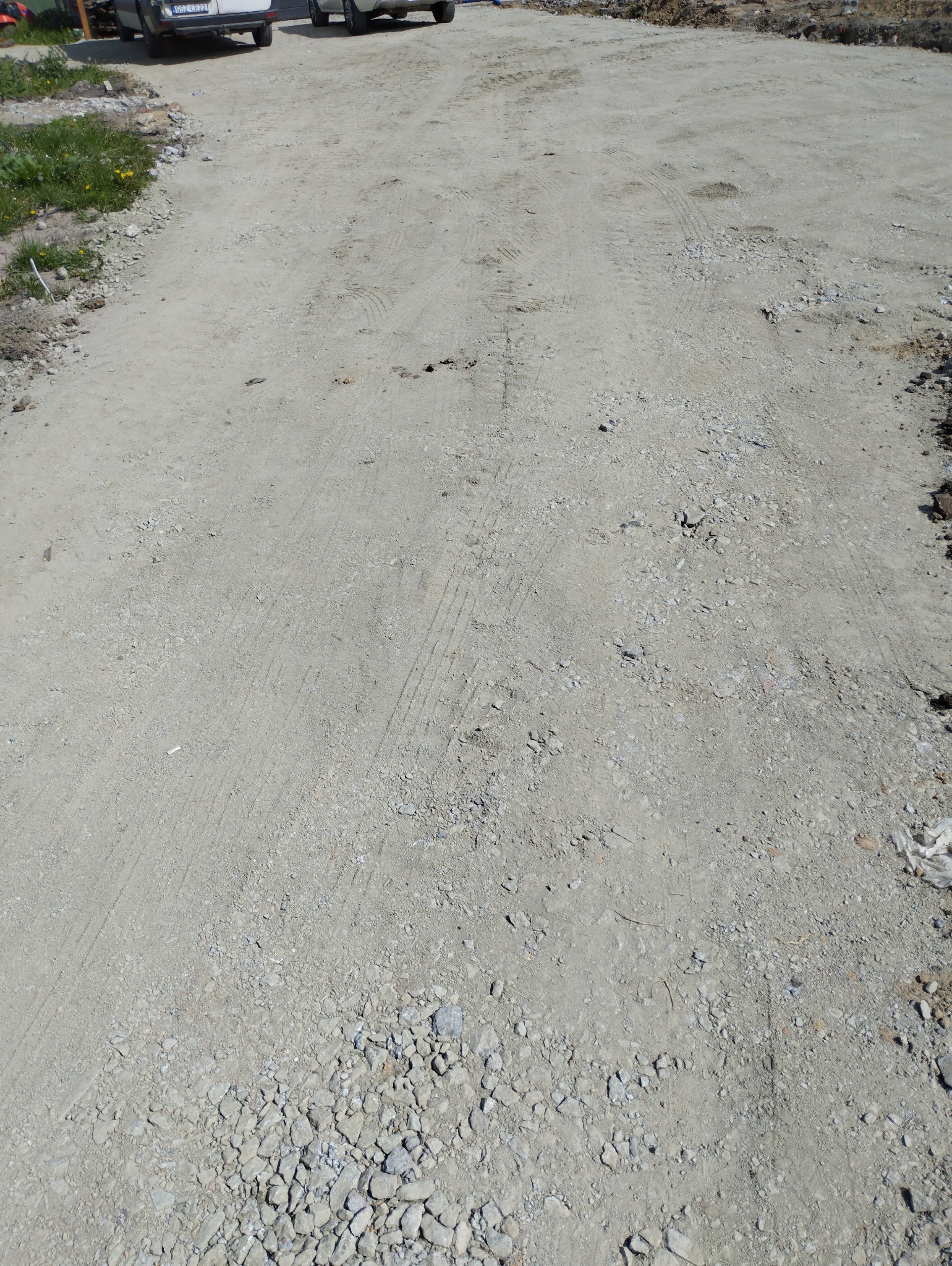 Niejednolita, szara nawierzchnia drogi gruntowej z widocznymi śladami opon samochodowych i luźnymi kamieniami, częściowo porośnięta trawą, w tle zaparkowane samochody.