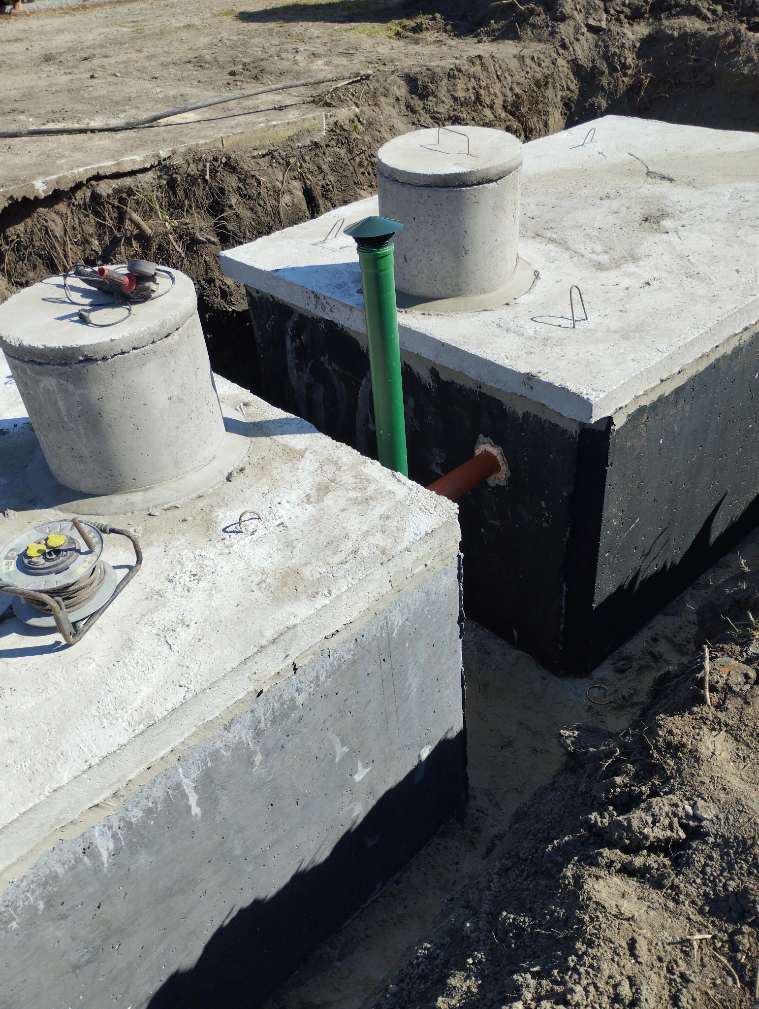 Dwa betonowe zbiorniki w wykopie, jeden z zaizolowaną ścianą zewnętrzną, połączone zieloną rurą, z narzędziami budowlanymi na górze jednego zbiornika.