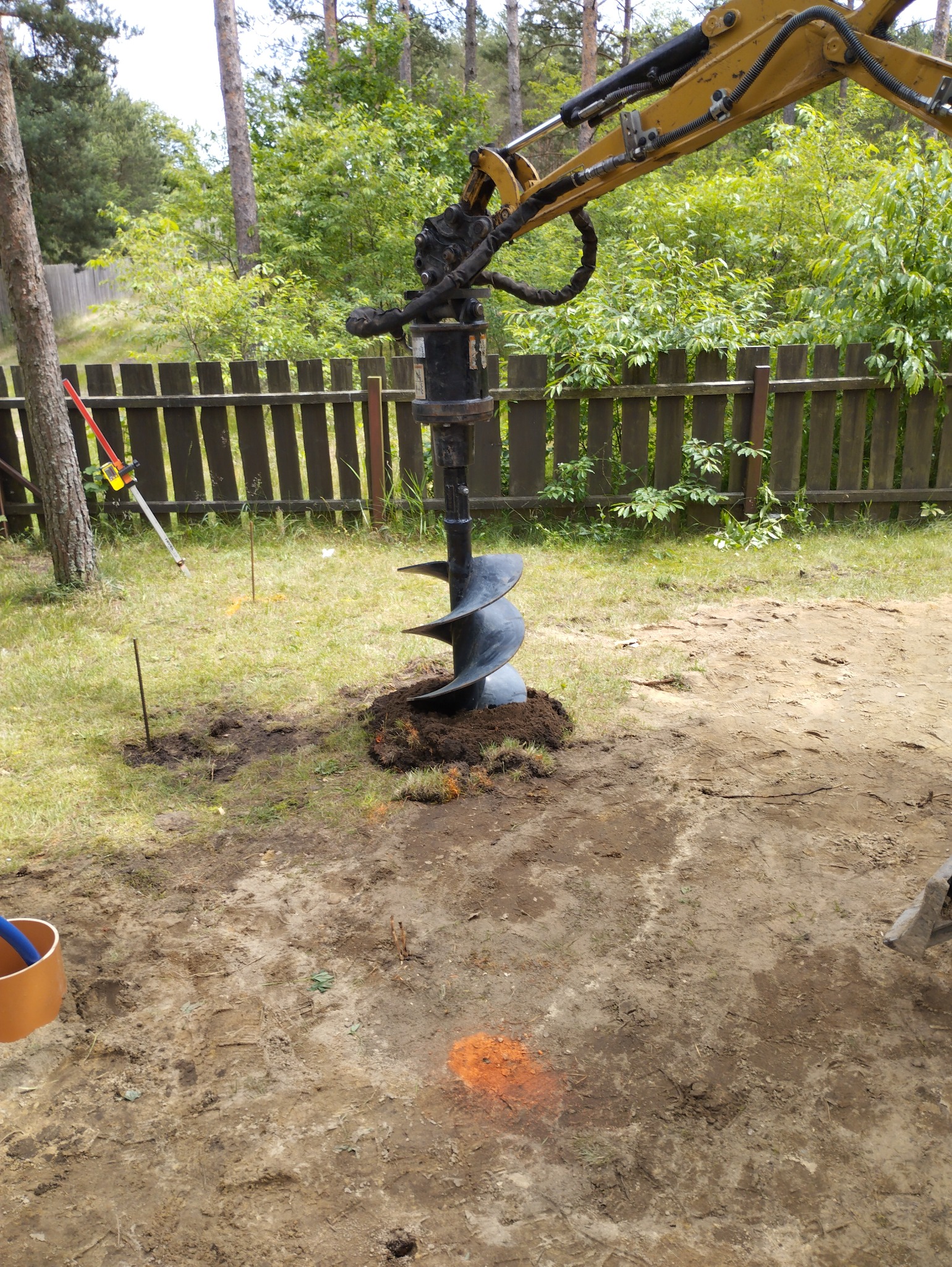 Żółta koparka z wiertłem świdrowym w trakcie wiercenia otworu w ziemi na trawiastym terenie, widoczne oznaczenia geodezyjne i drewniane ogrodzenie w tle.