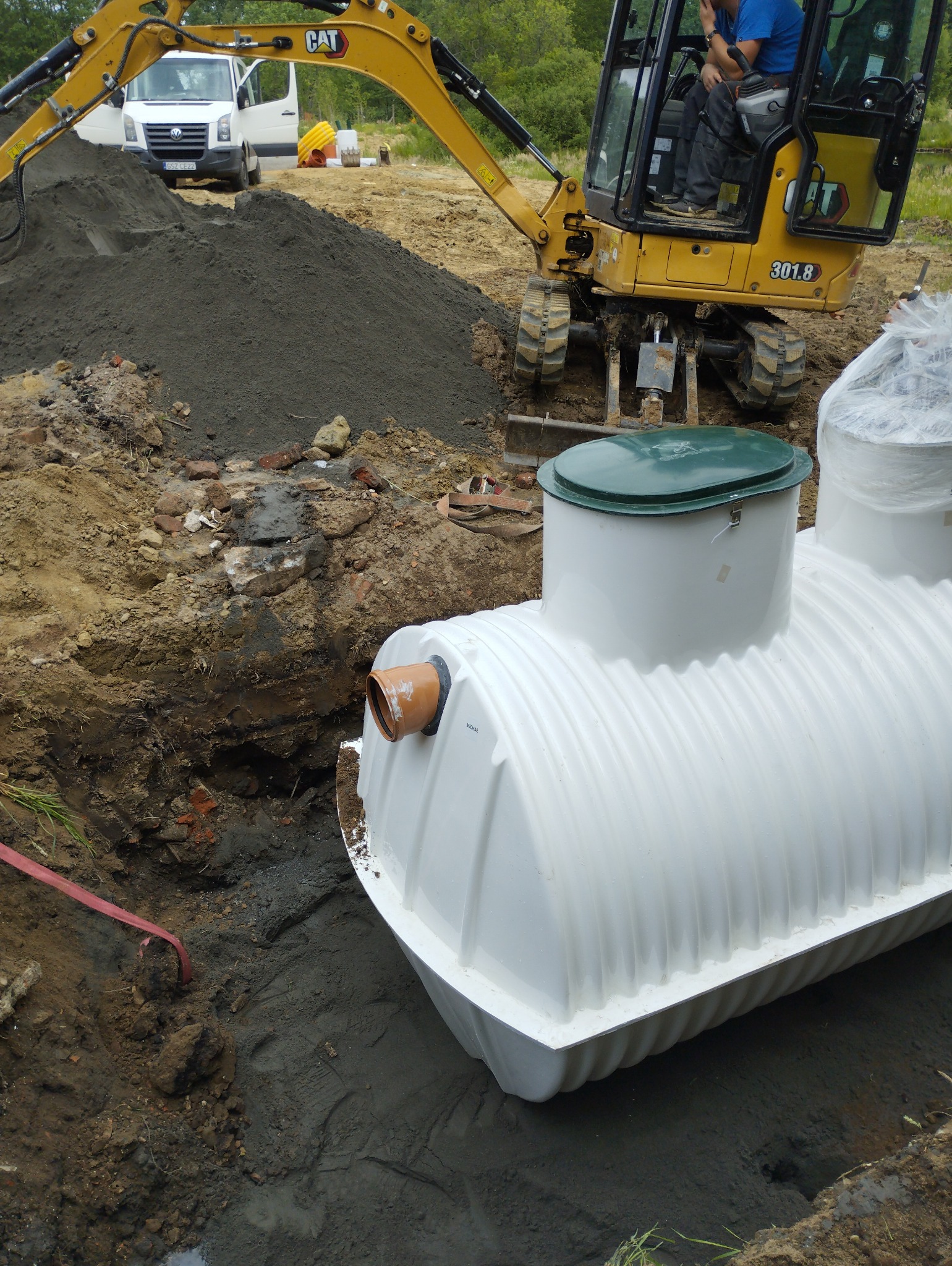 Biały zbiornik z zieloną pokrywą umieszczony w wykopie, widoczna koparka CAT 301.8 i sterta ziemi, w tle biały bus.