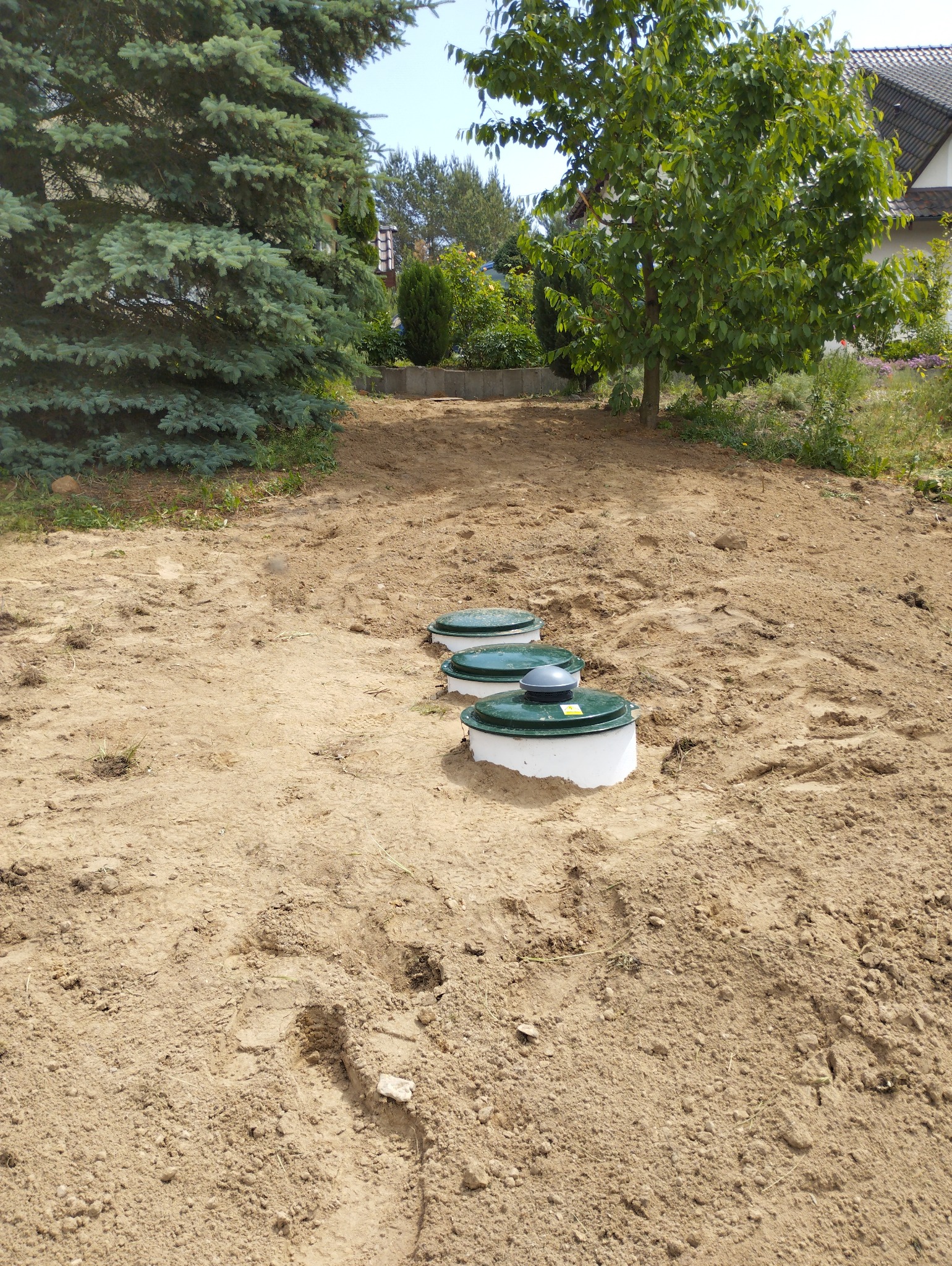 Trzy zielone pokrywy włazów z białymi podstawami, równo ustawione w piaszczystym gruncie, otoczone drzewami i roślinnością, z domem w tle.