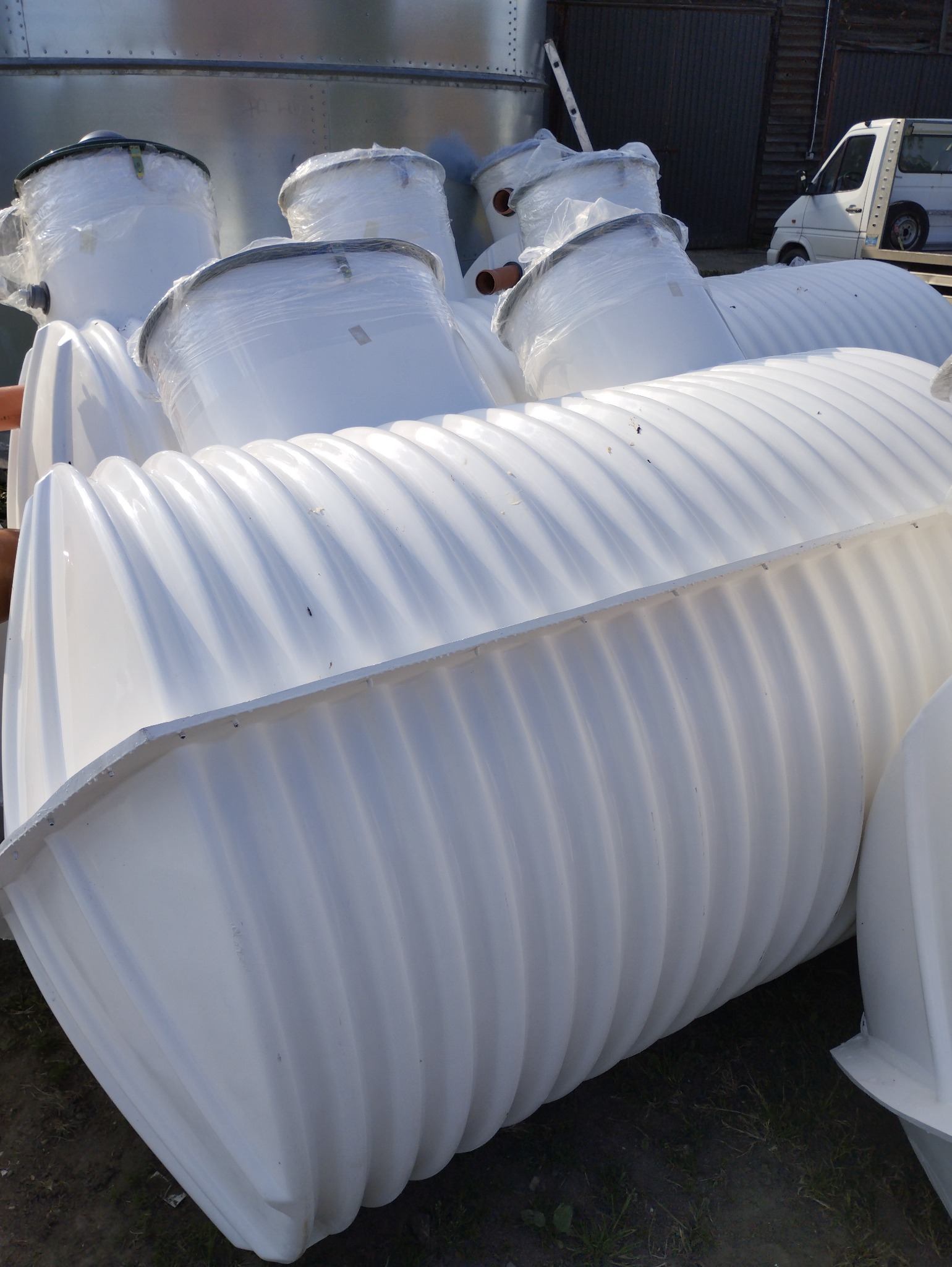 Stos białych, karbowanych zbiorników z tworzywa sztucznego, niektóre owinięte folią, na zewnątrz, z blachy falistej w tle.