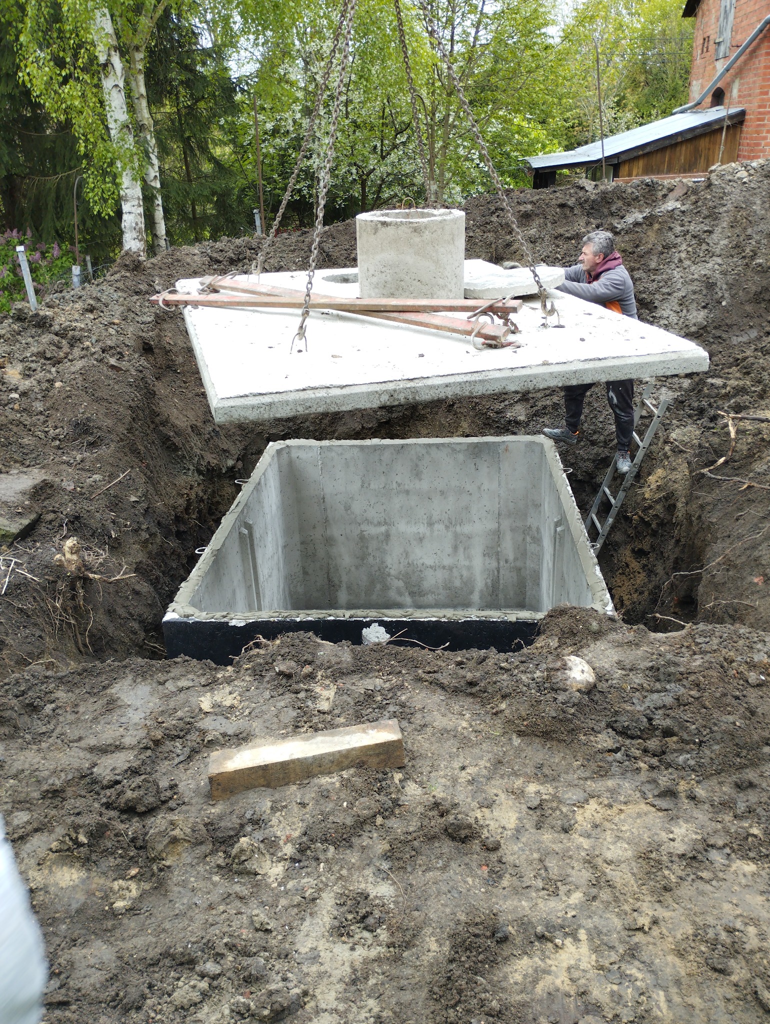 Montaż betonowego szamba: Robotnik na drabinie pomaga w opuszczeniu betonowej pokrywy na zbiornik, widoczne łańcuchy dźwigu, wykop ziemny i otaczająca zieleń.