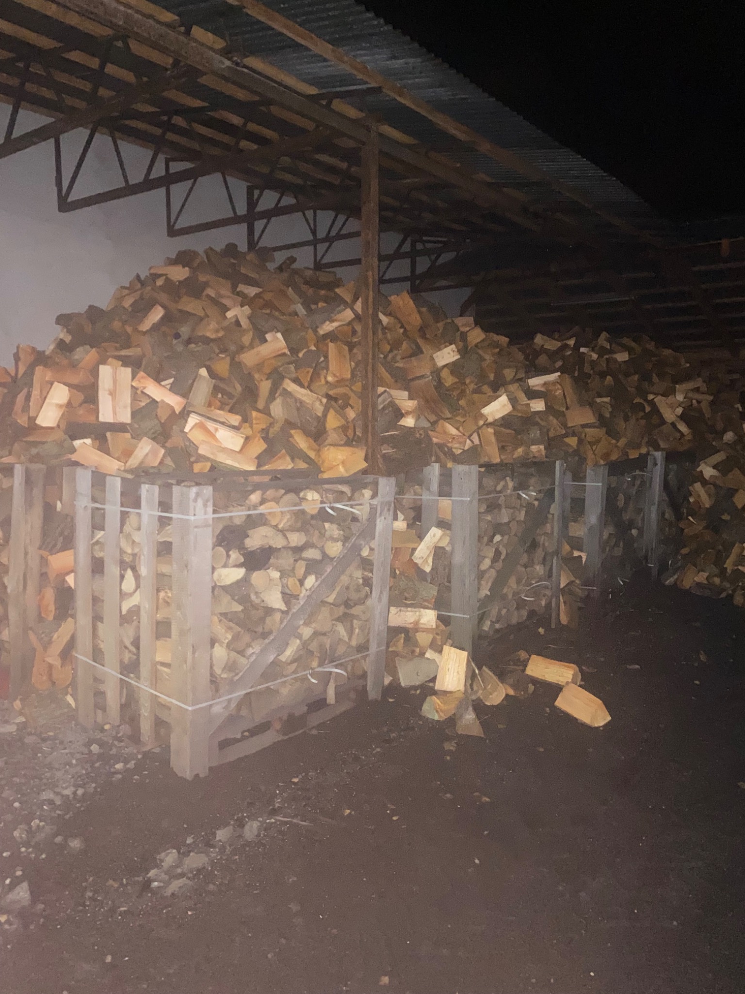 Stos pociętego drewna opałowego w drewnianych skrzyniach, składowany pod zadaszeniem o konstrukcji stalowej.