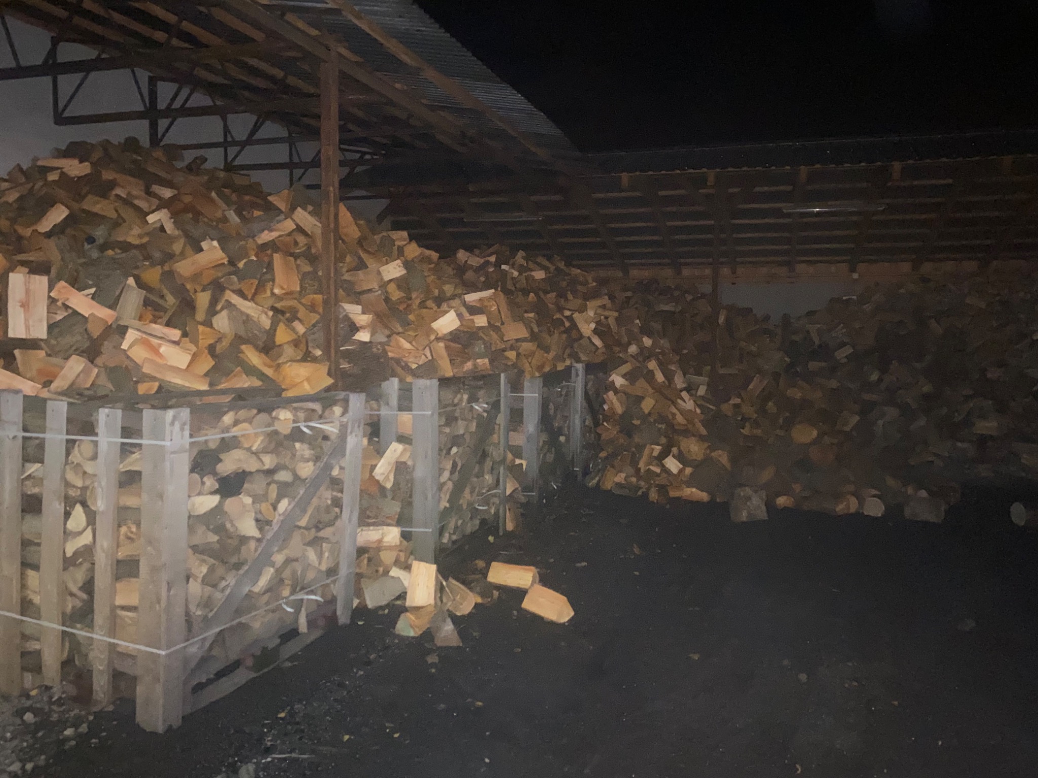 Stos pociętego drewna ułożony pod zadaszeniem, widoczne drewniane skrzynie wspierające konstrukcję, ciemne otoczenie.