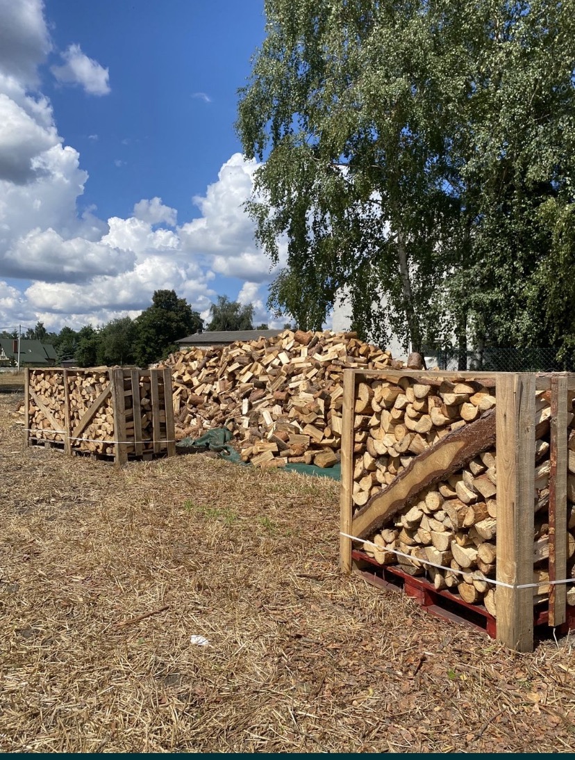 Stosy pociętego drewna ułożone w drewnianych skrzyniach na paletach, z dużym stosem drewna w tle i drzewem liściastym oraz błękitnym niebem z chmurami w tle.