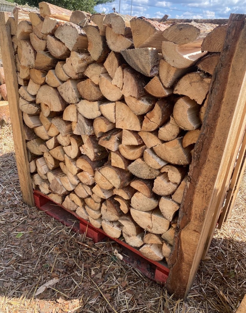 Rzędami ułożone, pocięte kawałki drewna w drewnianej skrzyni na czerwonej palecie, widoczne na tle ściółki leśnej.