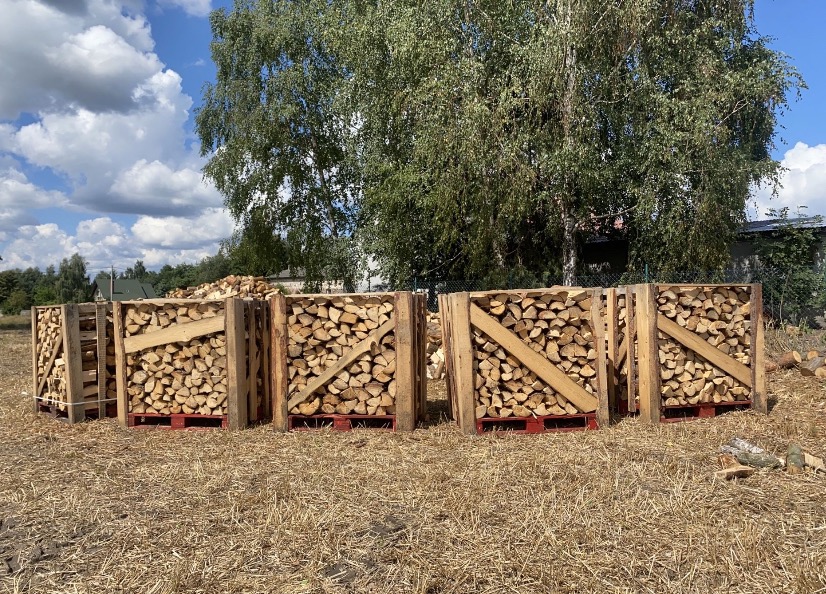 Ułożone drewno w skrzyniach na paletach, wysuszone na polu w słoneczny dzień, drzewo w tle.