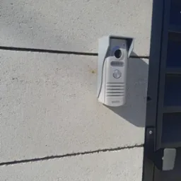 Szary wideofon marki Eura zamontowany na betonowym słupku ogrodzeniowym, widoczne elementy furtki.