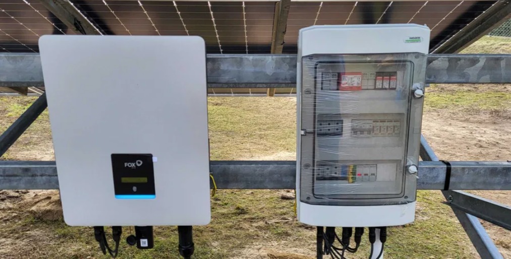 Biała skrzynka elektryczna z przełącznikami i okablowaniem, obok falownik FOX ESS zamontowane na konstrukcji wsporczej paneli fotowoltaicznych, zabezpieczona folią ochronną.