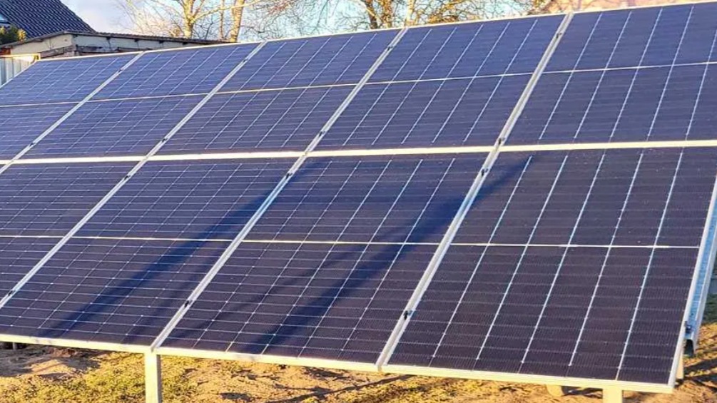 Zestaw paneli fotowoltaicznych na metalowej konstrukcji, zainstalowany na trawiastym terenie przy budynku z ciemnym dachem, widoczne cienie padające na panele.