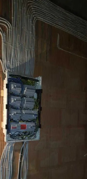 Rozdzielnica elektryczna z licznymi przewodami ułożonymi w wiązki, zamontowana na surowej ścianie z cegły ceramicznej.