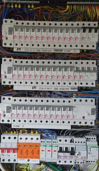 Szczegółowe ujęcie rozdzielnicy elektrycznej z trzema rzędami wyłączników ETI oraz zabezpieczeniami przeciwprzepięciowymi, widoczne okablowanie w różnych kolorach.