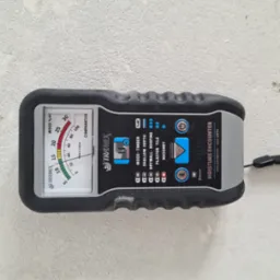 Wilgotnościomierz Tramex Moisture Encounter, ustawiony na pomiar wilgotności w murze, leży na szarej, szorstkiej ścianie.