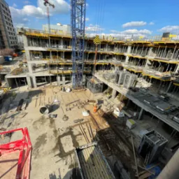 Wysoki kąt widzenia na plac budowy wielopiętrowego budynku mieszkalnego w trakcie wznoszenia, z widocznymi żurawiami wieżowymi, szalunkami, zbrojeniami i pracownikami w odblaskowych kamizelkach.