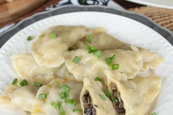 Porcja ręcznie robionych pierogów z farszem grzybowym, posypanych świeżym szczypiorkiem, ułożonych na białym talerzu z ozdobnym brzegiem.