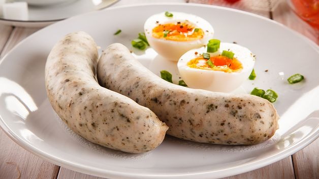 Dwie białe kiełbaski i dwa przekrojone na pół jajka na twardo, posypane szczypiorkiem, ułożone na białym talerzu na jasnym drewnianym stole.