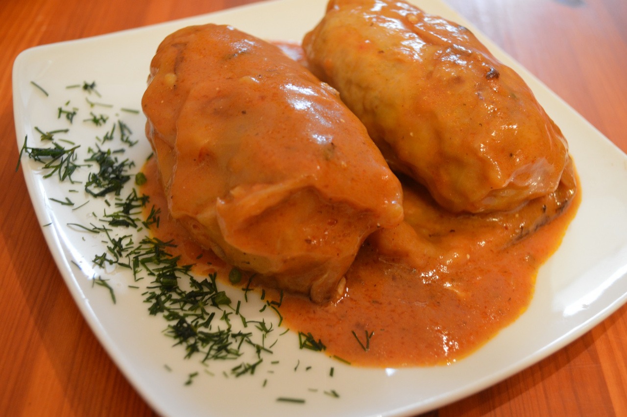 Dwa gołąbki w pomidorowym sosie na białym kwadratowym talerzu, udekorowane świeżym koperkiem, ustawione na drewnianym stole.