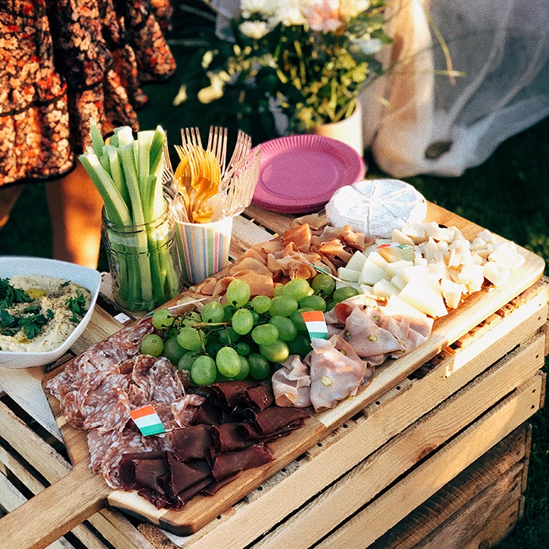 Deska serów, wędlin i owoców z flagami, podana na drewnianej skrzynce na trawie, obok seler naciowy w słoiku i dip.