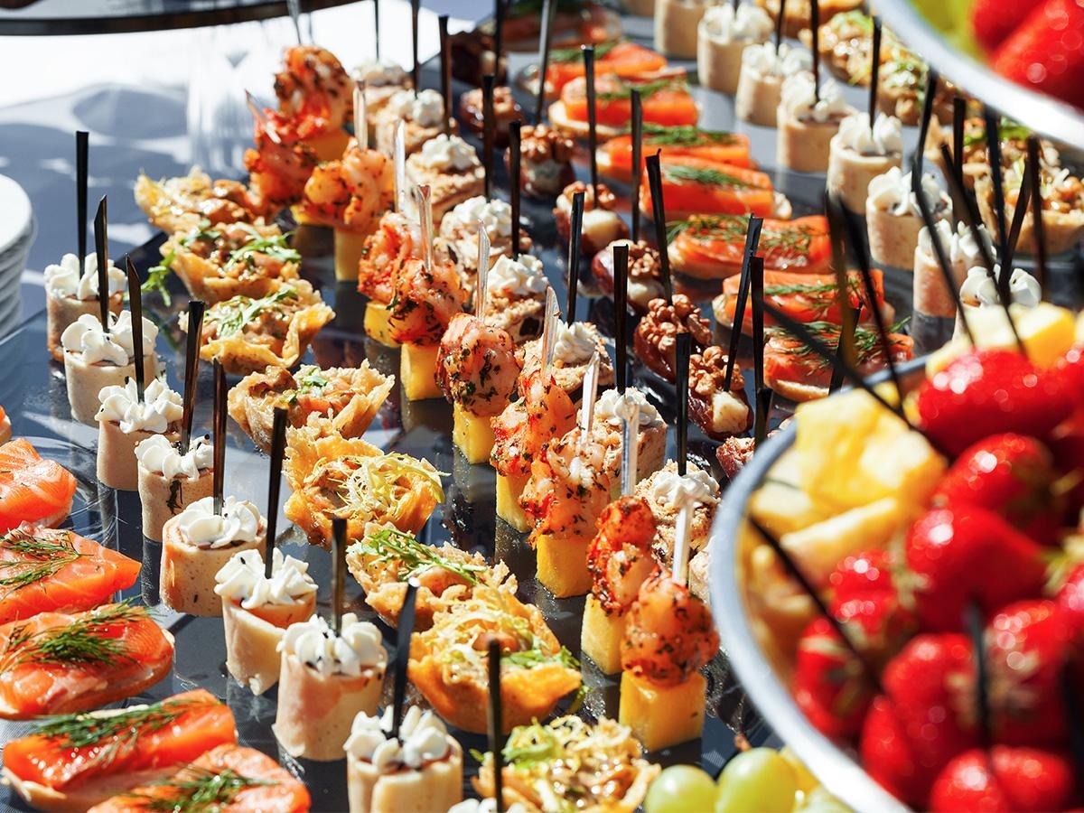 Elegancko ułożone, różnorodne przekąski na przyjęciu: kanapeczki z łososiem, mini tartaletki, szaszłyki z krewetkami i ananasem, oraz miska świeżych truskawek, wszystko na szklanym stole.
