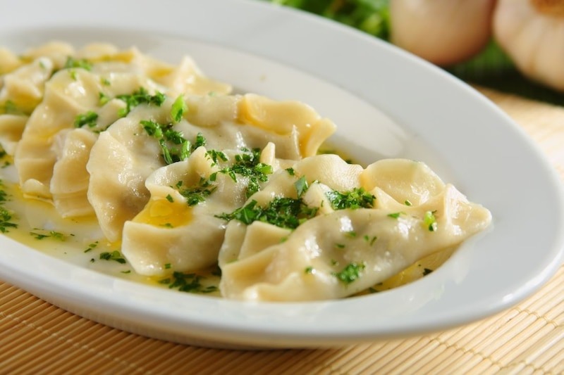 Ręcznie lepione pierogi okraszone masłem i posypane świeżą natką pietruszki, podane na białym talerzu, widoczne czosnek i zieleń w tle.