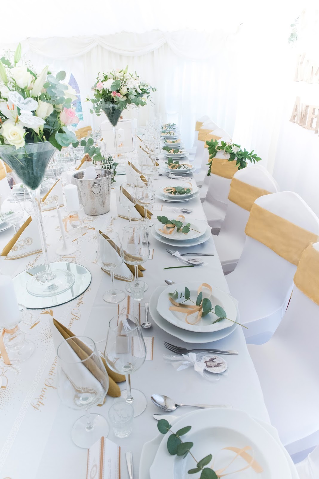 Elegancko nakryty stół z białym obrusem, złotymi akcentami i kwiatowymi dekoracjami, przygotowany na uroczystość. Widoczne białe krzesła z ozdobnymi złotymi wstęgami.