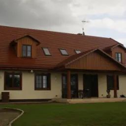 Dom jednorodzinny z brązowym dachem z dachówki ceramicznej, drewnianą podbitką i oknami. Widoczne okna dachowe i antena. Elewacja w kolorze kremowym.
