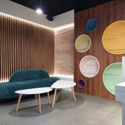 Blenda Studio meble na wymiar - Nowoczesne wnętrze z zieloną kanapą, białymi stolikami kawowymi i ścianą z drewnianymi lamelami oraz kolorowymi półkami w kształcie okręgów.