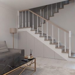 Blenda Studio meble na wymiar - Nowoczesne schody z białymi balustradami i drewnianymi stopniami, zintegrowane z szarymi szafkami pod schodami w salonie z szarą sofą i marmurowym stolikiem kawowym.