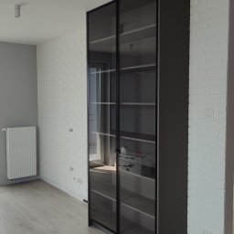Blenda Studio meble na wymiar - Nowoczesna, wysoka szafa z przesuwnymi, szklanymi drzwiami w ciemnej ramie, na tle białej ściany z cegły i jasnej podłogi. Minimalistyczny design.