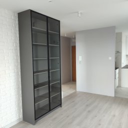 Blenda Studio meble na wymiar - Szara, przeszklona szafa z czarnymi ramami, stojąca na jasnej podłodze, na tle białej ściany z cegły. Widoczne wnętrze mieszkania z elementami kuchni.