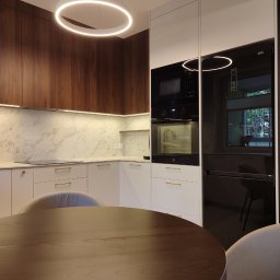 Blenda Studio meble na wymiar - Nowoczesna kuchnia z drewnianymi i białymi szafkami, marmurowym blatem i okrągłym oświetleniem. Widoczny stół z krzesłami. Minimalistyczny design.